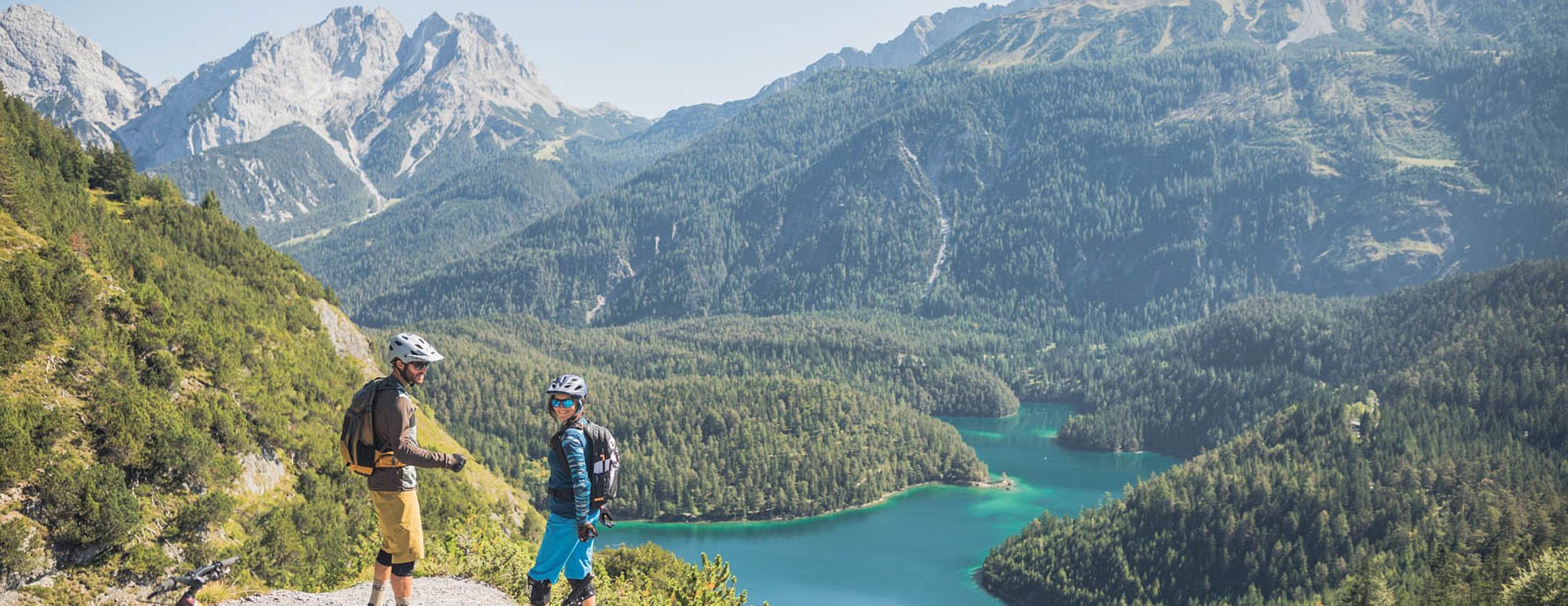 Zwei Mountainbiker genießen Aussicht auf türkisfarbenen See und bewaldete Alpenlandschaft