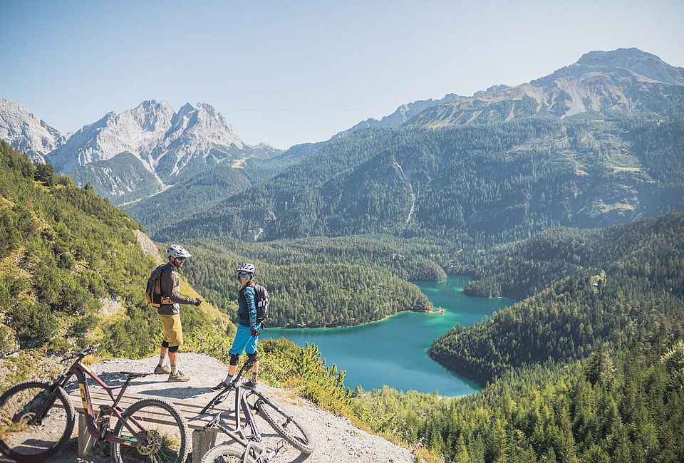 Zwei Mountainbiker genießen Aussicht auf türkisfarbenen See und bewaldete Alpenlandschaft