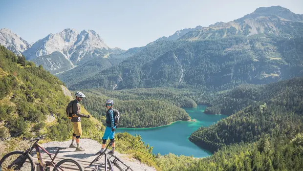Zwei Mountainbiker genießen Aussicht auf türkisfarbenen See und bewaldete Alpenlandschaft