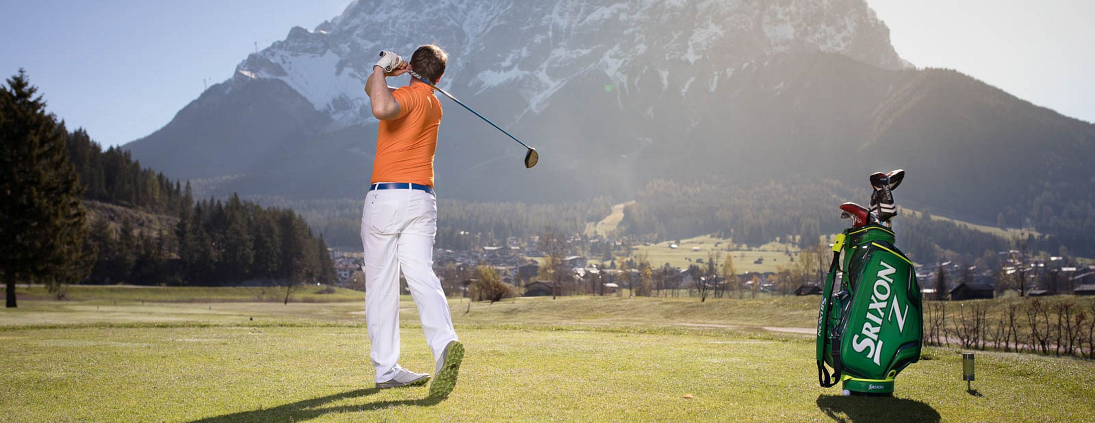 Man tees off on a golf course with a view of snowy mountains and a sunny sky