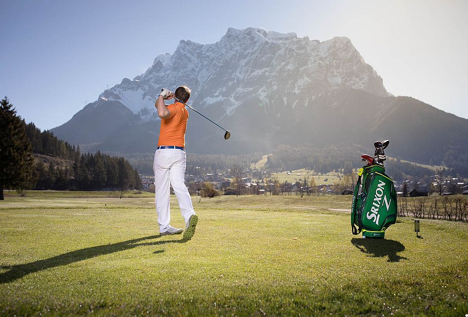 Man tees off on a golf course with a view of snowy mountains and a sunny sky