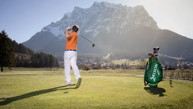 Mann beim Abschlag auf Golfplatz mit Blick auf verschneite Bergkulisse und sonnigem Himmel